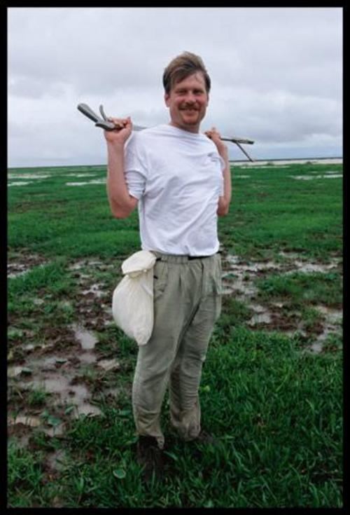 Joseph Bruno Slowinski smiling while holding a snake tong and wearing a white t-shirt, gray pants, and a white cloth bag tied around his waist