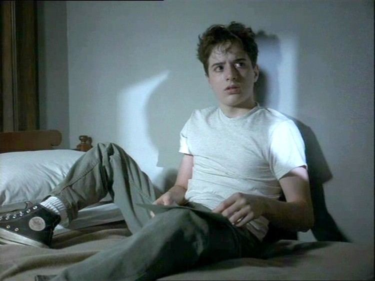 Joseph Perrino sitting on the bed, holding a paper, and looking at something with a serious face in a scene from the 1996 American legal crime drama film, Sleepers. He is wearing black and white sneakers, white socks, a white t-shirt, and gray pants.