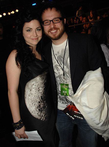 Amy Lee and Josh Hartzler are smiling while Amy wearing a black sleeveless blouse and her husband is wearing a black jacket and white t-shirt