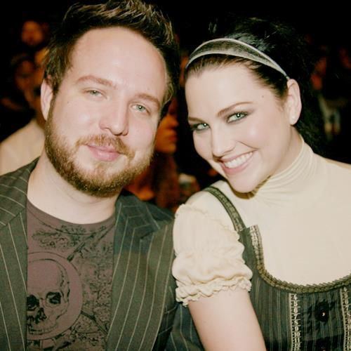 Josh Hartzler and Amy lee are smiling while Josh is wearing a striped coat and t-shirt and his wife is wearing a cream blouse underneath a sleeveless blouse