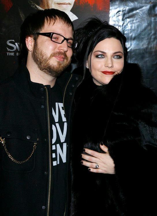 Josh Hartzler and Amy lee are smiling while Josh is wearing a black jacket, black t-shirt, and eyeglasses and his wife is wearing a black fur coat