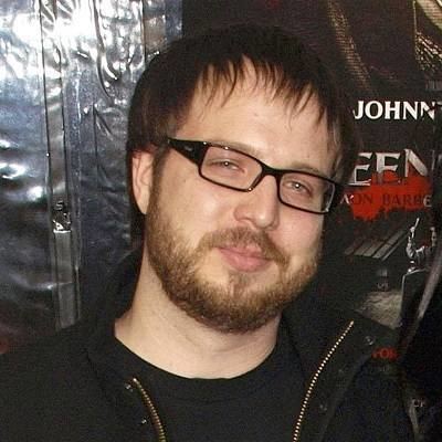 Josh Hartzler with a tight-lipped smile, mustache, and beard while wearing a black jacket, black t-shirt, and eyeglasses