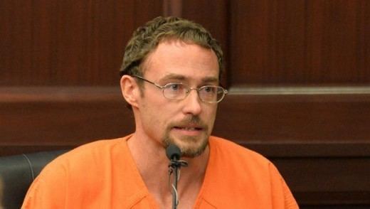Josh Phillips speaking, with mustache and beard, while wearing eyeglasses and orange t-shirt prison uniform