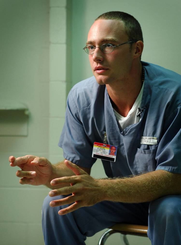 Josh Phillips, in one of his interviews, speaking with hand gestures, and wearing a white shirt underneath a blue shirt, eyeglasses, blue pants, and identification badge