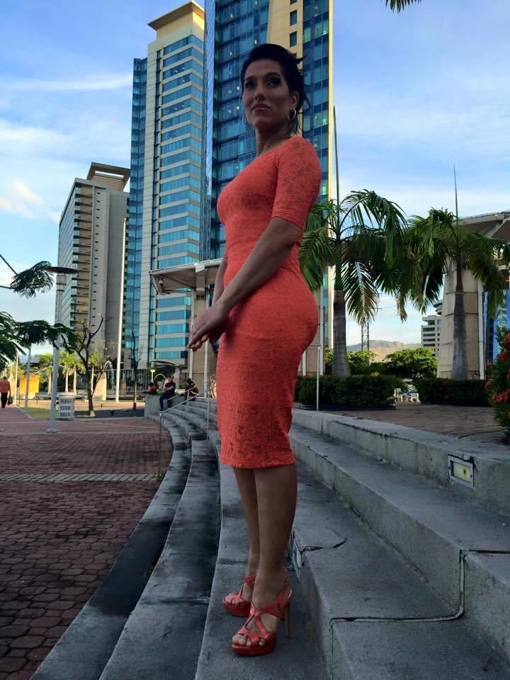 Jowelle de Souza smiling while standing on a step street with tall buildings, people, and trees in the background and wearing an orange lace dress, heels and round earrings