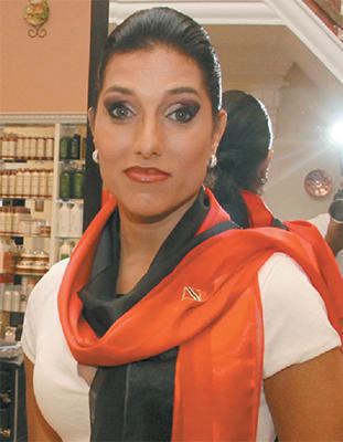 Jowelle de Souza smiling with a mirror in the background, wearing a white blouse, a black and orange scarf around her neck with the printed letter "N" and silver earrings