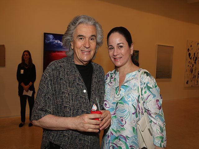Inside the art gallery is a woman (left) standing, serious, has long black hair, her left hand holding her right hand while holding a paper, wears a necklace, a black watch on her left hand, a black long sleeve top with white pocket, black pants and shoes. In the middle is Juan Ruiz Healy smiling, has white hair, both hands holding a glass bottle in red color cloth, with a red-white stick inside, behind him from left to right are different kinds of paintings, he is wearing a black shirt under a black and white polo. A woman (right) is smiling, has black hair, wears silver earrings, a white bag on her left shoulder and a blue-colored long-sleeve top with different designs and colors.