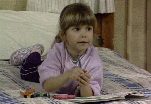 Judith Barsi as Anna lying in a swimmer's position on a bed while holding a crayon in a scene from Punky Brewster, a 1984 American sitcom television series about a young girl being raised by a foster parent. Judith is wearing grey shoes, a purple sweatshirt, and black pants.