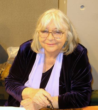 Judy Cornwell smiling while wearing a dark violet long sleeve blouse, white scarf, and eyeglasses