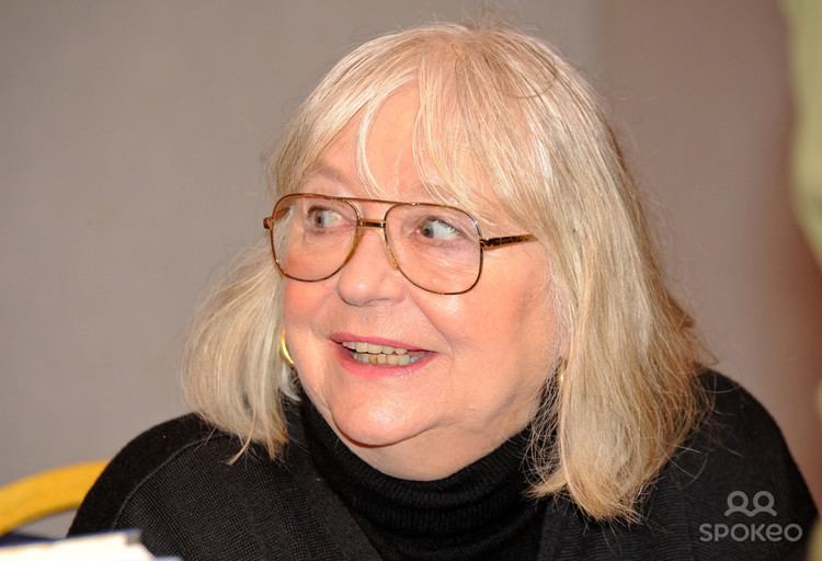 Judy Cornwell smiling while looking at something and wearing eyeglasses and a black blouse