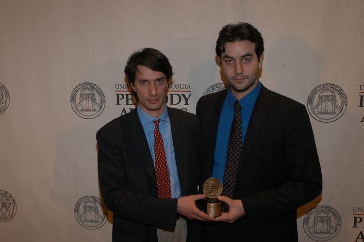 Gédéon Naudet (left) and Jules Naudet (right), posing with their Peabody Award while wearing a black coat, blue long sleeves, and polka dot necktie