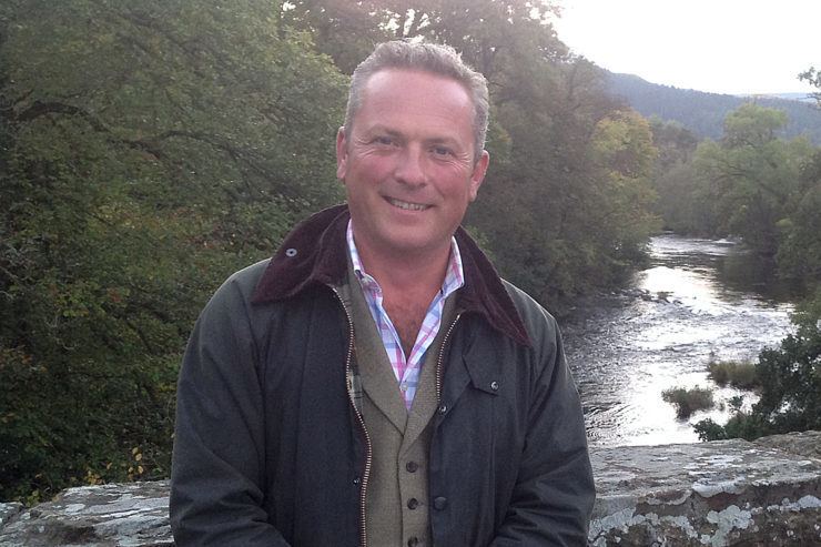 Jules Hudson smiling while leaning on the bridge on the top of a river, wearing a jacket, over a brown vest, and checkered multi-colored polo shirt.