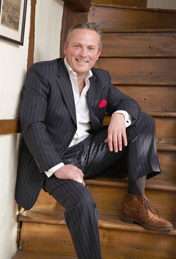 Jules Hudson smiling while sitting on the stairs, wearing a black striped coat and pants, white long sleeves, black socks, and brown shoes.