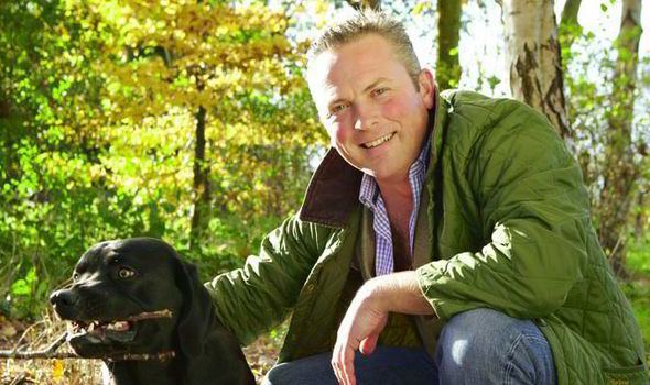 Jules Hudson smiling while holding a black dog biting a branch of a tree, wearing a green jacket over a blue checkered polo shirt, and blue jeans.