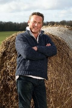Jules Hudson smiling beside hay while his arms crossed on his chest beside, wearing a gray sweatshirt over a white shirt, and black pants.