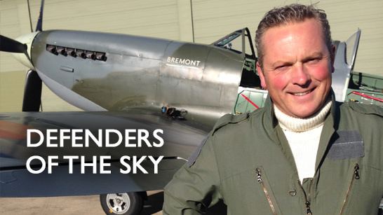 Jules Hudson smiling with a single-seat fighter-bomber on his back and wearing a green long sleeve coat over a white knitted shirt.