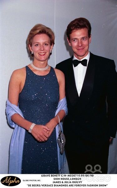 Julia and James Ogilvy are smiling. Julia with short blonde hair, wearing earrings, a necklace, a blue sleeveless dress with a purple scarf while James wearing a black coat over white long sleeves, a black bow tie, and black pants.
