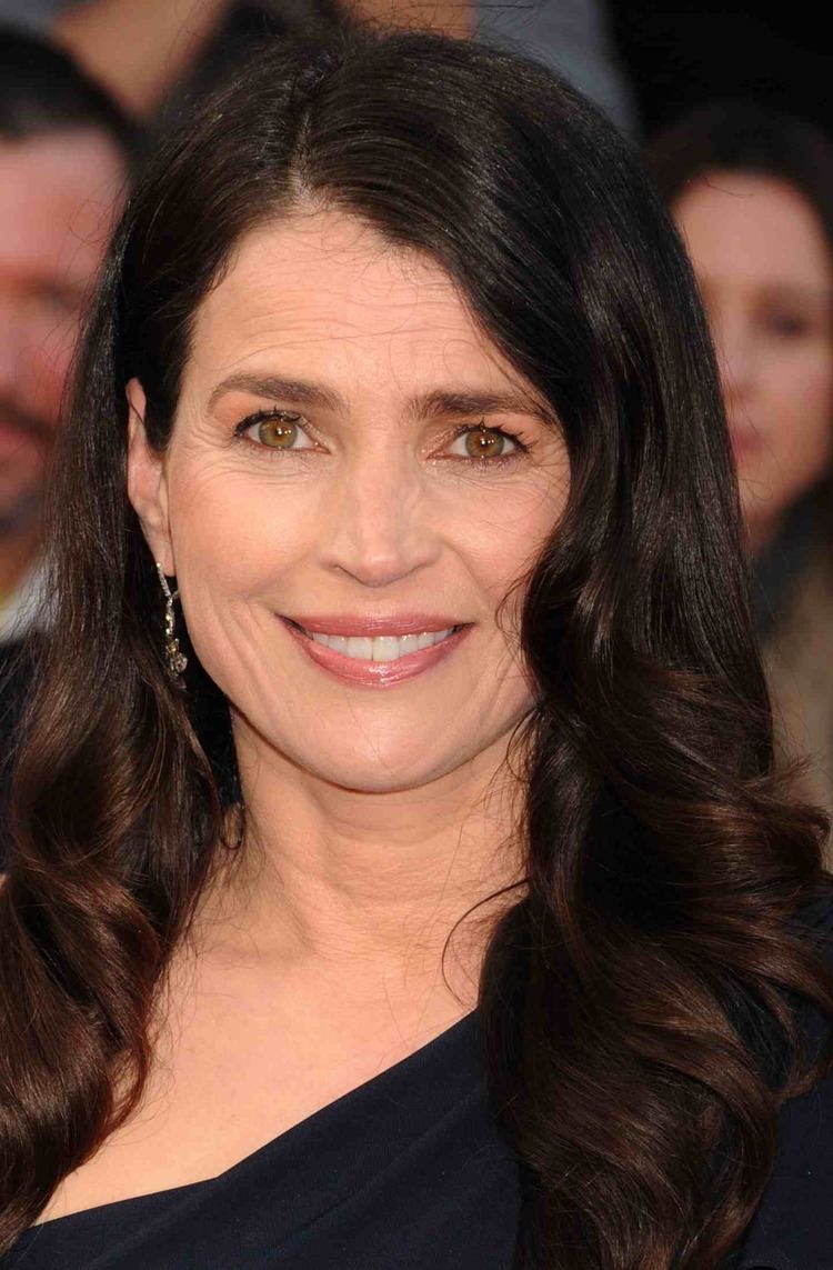 Julia Ormond smiling with curly hair and hazel eyes wearing dangled earrings and a one-sleeve dress.