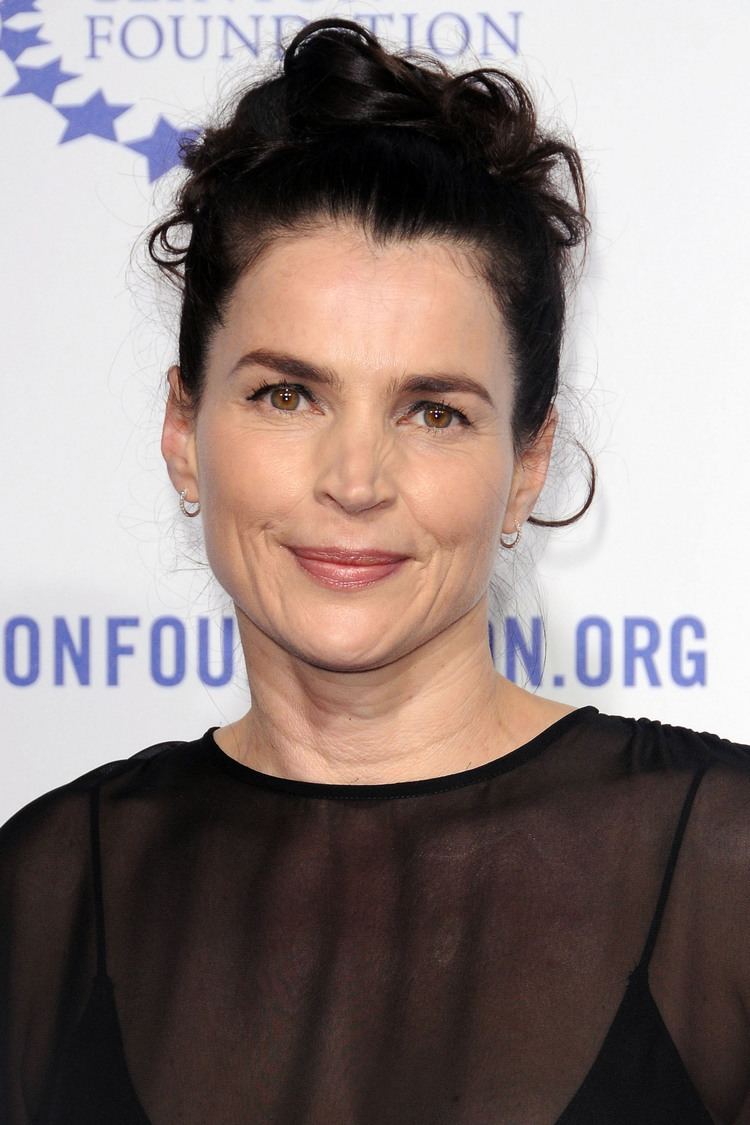 Julia Ormond smiling with her curly bun hairstyle and hazel eyes, wearing a small round earring and a black sleeveless under a see-through black top
