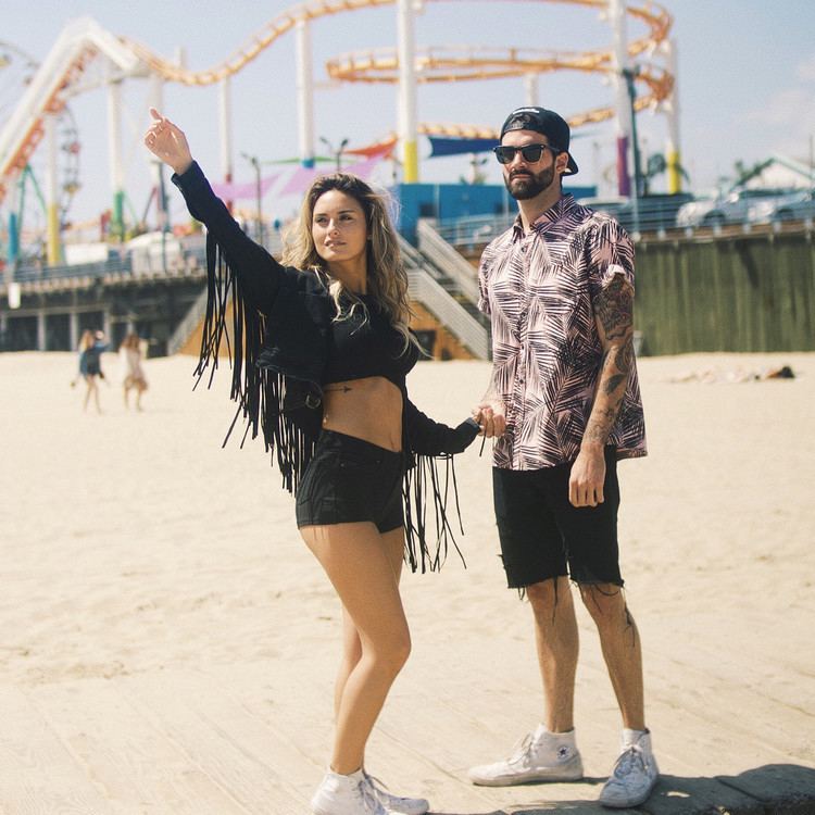 Julia Rose pointing her finger above with Stephen McHugh looking at it and holding her hand. Julia wearing a sexy black top with a black blazer and black shorts while Stephen with beard and mustache, wearing sunglasses, a black cap, a polo shirt, and black shorts.