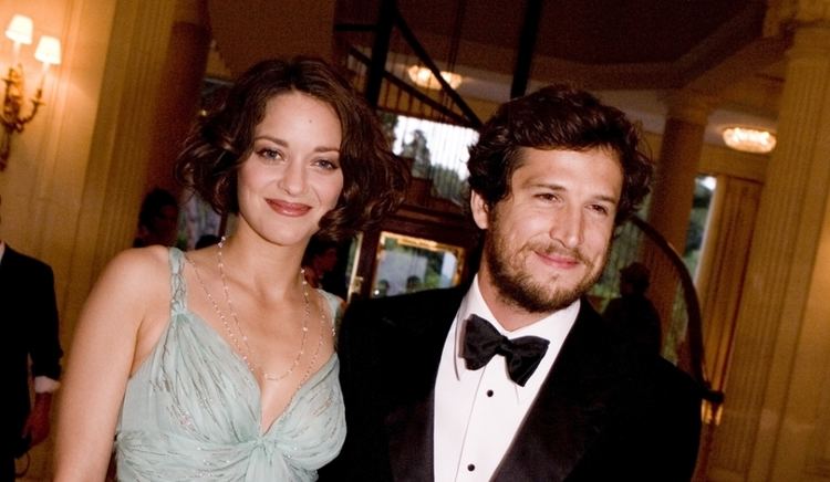 In a room with large pillars yellow wall lamps and spiral staircase at the back, from left, Marion Cotillard is smiling, has brown hair wearing a necklace and a gray sleeveless dress, at the right, Gauilaume Canet is smiling, standing, has brown hair a beard and a mustache wearing a white polo with black bowtie under a black coat.