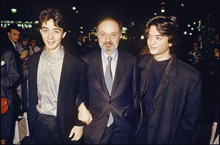 In a party outside an event with group of people, lights and buildings at the back, in front from left, Julien Rassam is smiling, standing hands down, has black hair wearing a gray polo under a black coat and a black pants, in the middle, Claude Berri is smiling, standing, hand closed with his right arm holding Julien, has black hair, white beard and mustache, wearing a white polo with silver necktie under a black coat and black pants, at the right, Thomas Langmann is smiling, standing looking to his right has black hair wearing a black shirt under a black jacket.