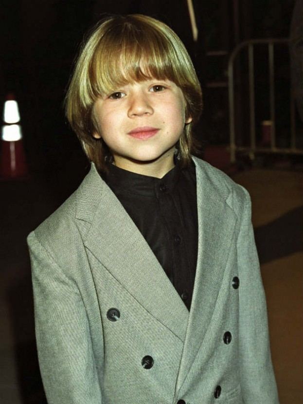 Justin Cooper smiling, with blonde hair and wearing a gray suit and a black shirt.