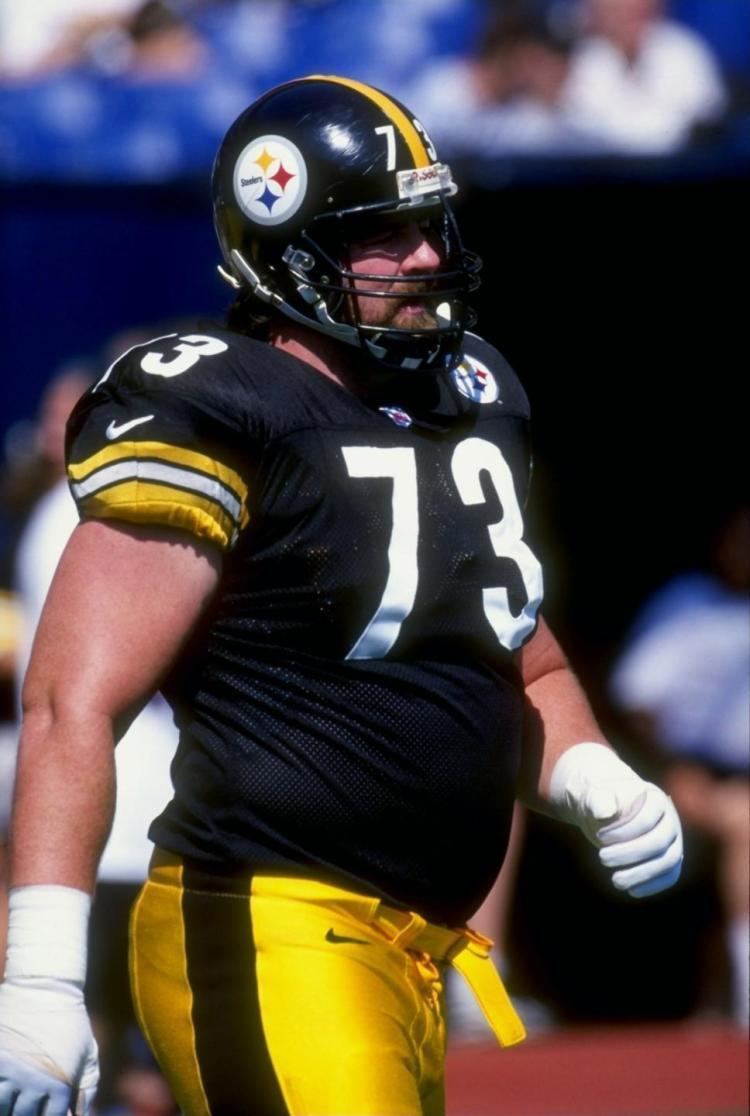 Justin Strzelczyk looking afar with mustache and beard while wearing a black and yellow football helmet, number seventy-three black and yellow jersey, black and yellow shorts, and white gloves