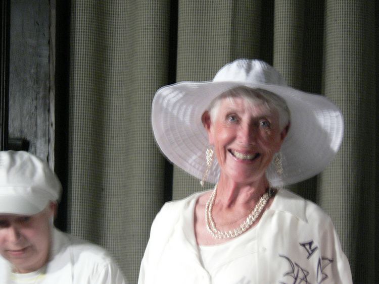 Justine Lord (right) is smiling, has white hair, wearing white earrings, a necklace, a hat, and a dress with a print on left. A woman (left) is serious, looking down, wearing a white cap and top.