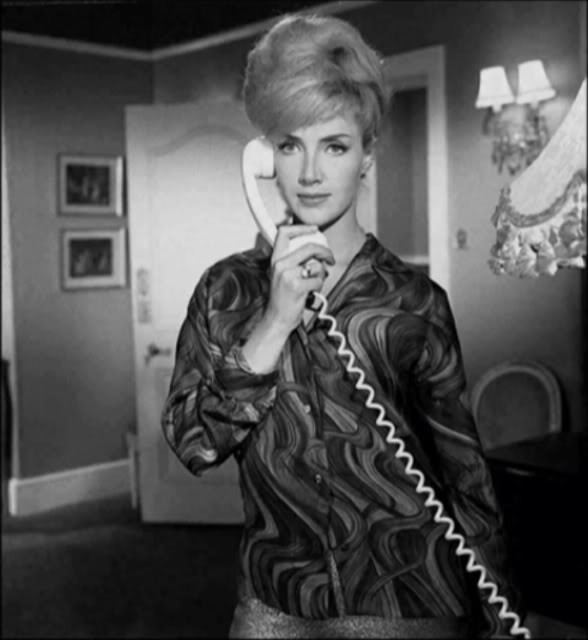 Justine Lord is serious, has long blonde hair, standing inside a room with frames on her right, and a lamp on her left, behind is a door, her right hand is holding a telephone, and she is wearing black earrings and black long sleeves with design.