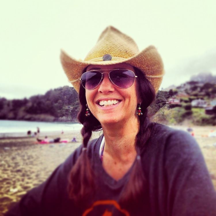 Justine Miceli smiling while wearing a brown hat, shades,and black t-shirt