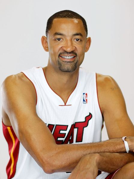 Juwan Howard smiling with a beard and mustache with crossed arms resting on top of his knee while wearing a red and white jersey with the word "HEAT" and the NBA logo written on it