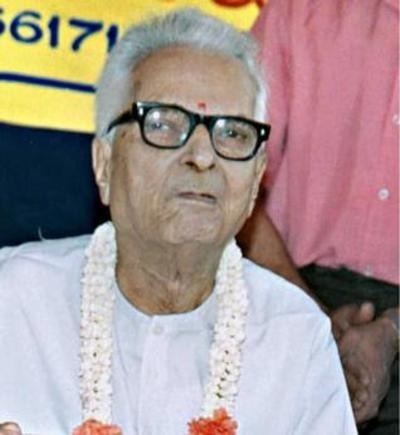 K. S. Narasimhaswamy smiling with a red mark on his forehead while wearing eyeglasses, white shirt, and garland
