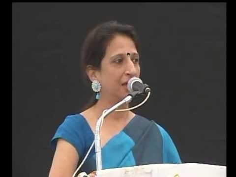 Kaajal Oza Vaidya wearing earrings, and a blue dress standing beside a podium with a microphone.