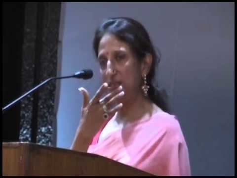 Kaajal Oza Vaidya wearing earrings, rings, and a pink dress standing beside a podium with a microphone.