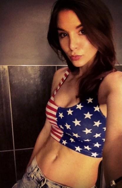 Kacey Clarke pouting and holding the phone with her straight hair inside the comfort room wearing a bracelet on her left arm, an American flag sleeveless crop top, and jeans.