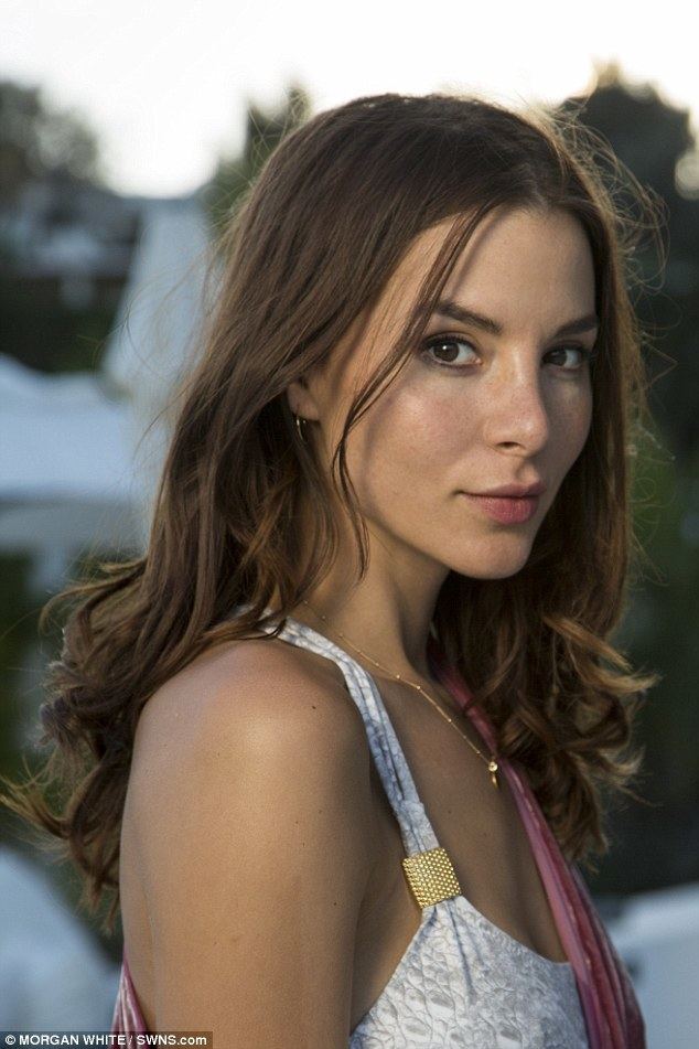 Kacey Clarke smiling with her curly brown hair wearing a small round earring, a gold necklace, and a sleeveless dress.