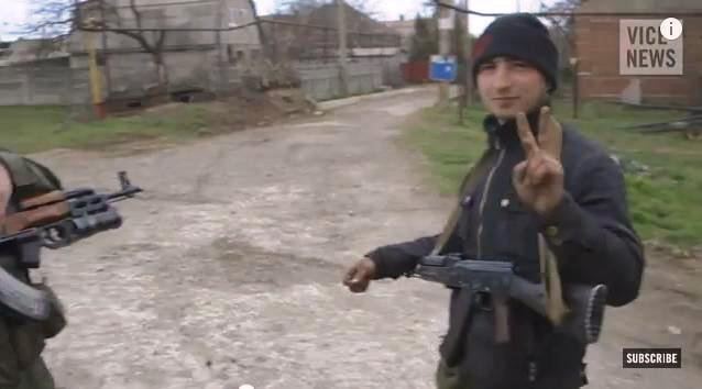 An armed Kadyrovtsy man smiling and showing a V hand gesture and wearing a black suit and bonnet