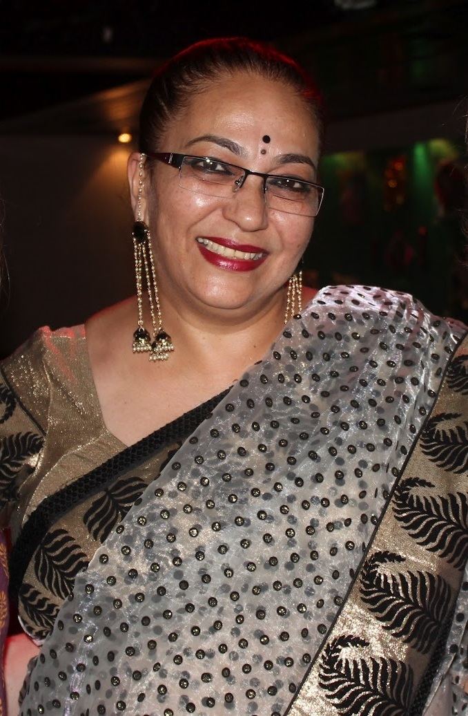Kalpana Iyer Here smiling with a black bindi and mole on her forehead while wearing earrings, eyeglasses, and a black and brown saree, and black and white polka dot see-through dupatta