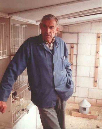 Karel Meulemans with a serious face while his hand on his pocket inside a pigeon's cage, wearing a blue jacket, checkered polo shirt, and gray pants.