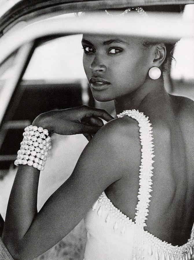 Karen Alexander posing inside the car while wearing a beaded sleeveless top, pearl bracelet, and earrings