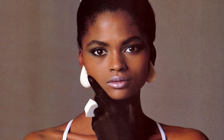Karen Alexander posing while hand on her chin and wearing a white top, white earrings, white ring, and black gloves
