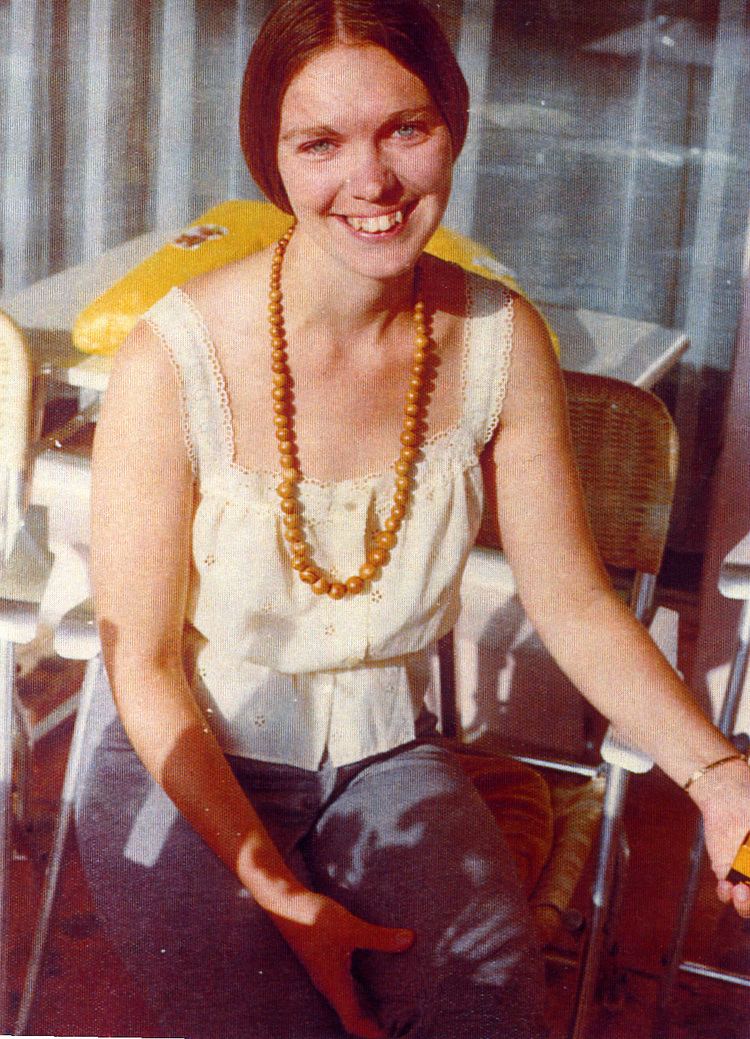A younger Karen Zerby Karen sitting down and smiling with reddish hair and wearing a white sleeveless blouse and a wooden necklace.