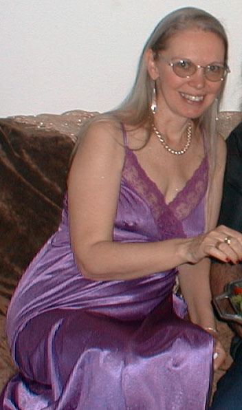 Karen Zerby smiling and sitting in a couch while wearing a purple sleeveless dress along with jewelry and eyeglasses.