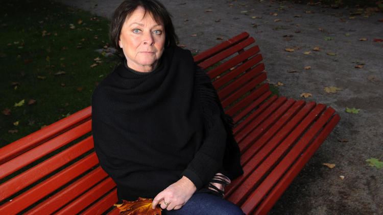 Kari Storækre sitting on a red bench and holding a dried leaf with a serious face and black short hair while wearing a ring, blue denim pants, and a black long sleeve blouse under a black shawl