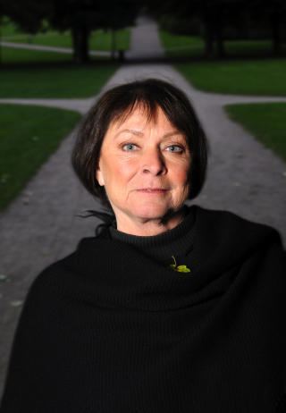 Kari Storækre with a tight-lipped smile, black short hair, and a leaf on her chest while wearing a black long sleeve blouse under a black shawl