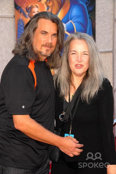 Robby Benson with a tight-lipped smile while Karla DeVito is smiling with closed eyes. Bobby with long hair, with a beard and mustache, wearing a black and orange shirt and black pants while Karla is with long blonde hair, wearing a necklace, a black sling bag, an ID and black dress.