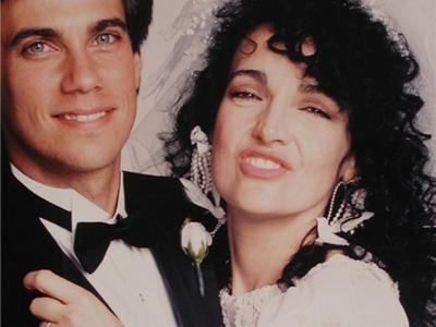 Robby Benson and Karla DeVito are smiling at their wedding. Robby is wearing a black coat with a white rose over white long sleeves and a black bowtie while Karla is with curly hair, wearing white earrings and a white wedding gown.