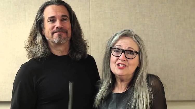 Robby Benson and Karla DeVito are smiling. Bobby with long hair, with a beard and mustache, wearing a black long sleeve shirt while Karla is with long blonde hair, wearing eyeglasses and a black blouse.