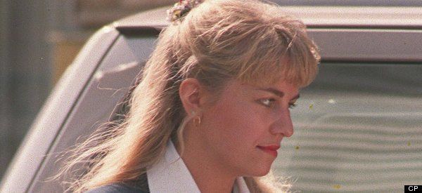 Karla Homolka is serious, walking in front of a gray car, looking to her left, has long blonde hair white tie, wearing gold earrings and white top under a blue-green coat.